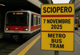 Cartello giallo con la scritta “Sciopero 7 novembre 2025 metro bus tram” alla stazione di Milano, con treno e tram ATM fermi sullo sfondo.